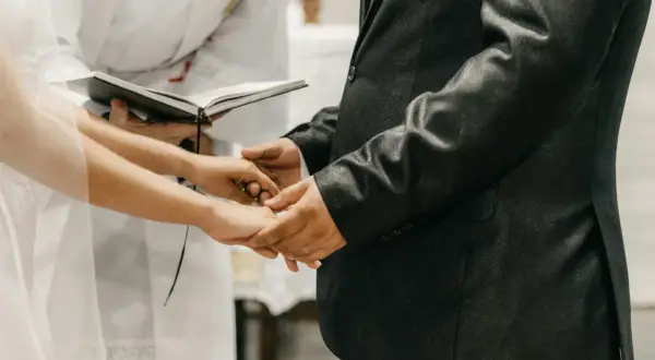 Mani che si stringono durante la dichiarazione matrimonio, con un libro aperto e un sacerdote sullo sfondo.