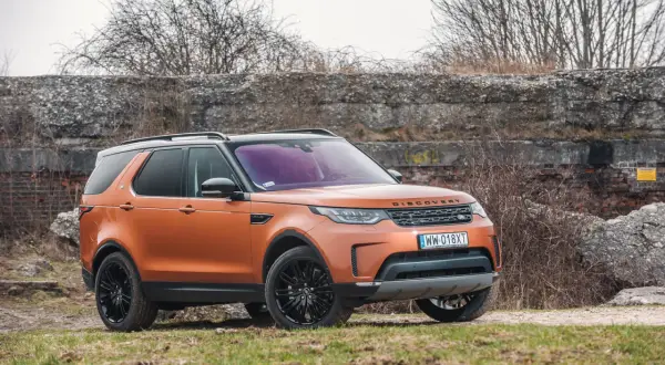 Pomarańczowy Land Rover Discovery 5 z czarnymi felgami stoi na trawiastym podłożu, na tle kamiennego muru i suchych krzewów.