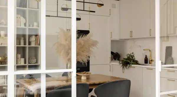 Elegancka kuchnia z białymi frontami i uchwytami w kolorze mosiądz. W centralnym punkcie stół z suszonymi trawami i czarnymi krzesłami.