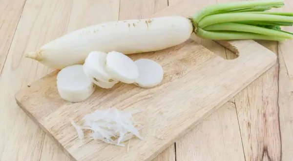 Ein weißer Rettich liegt auf einem Holzbrett, daneben liegen Scheiben und geraspelte Stücke. Perfekt, um Rettich zu fermentieren.