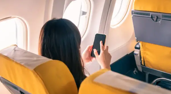 Kobieta w samolocie trzyma telefon, zastanawiając się, **co to tryb samolotowy**. Widok zza jej pleców na żółte siedzenia i okno.