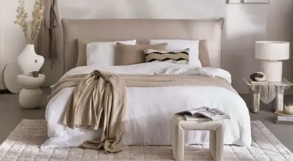 Ein gemütliches Schlafzimmer im shabby stil mit einem weißen Bett, beige Decke und Kissen. Ein Beistelltisch mit Lampe und Deko.