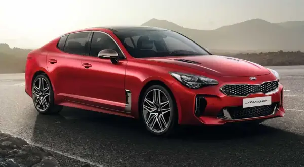 Kia Stinger CV rojo brillante, con llantas deportivas, aparcado en una carretera mojada con montañas al fondo.
