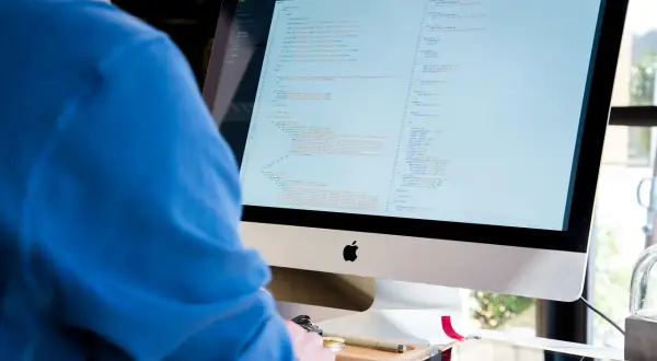 Programista IT w niebieskiej bluzie pochyla się nad monitorem z kodem.