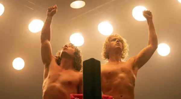 Zwei Wrestler mit blonden Locken, die unter Scheinwerfern die Fäuste gen Himmel strecken. Sie sind bereit, den "Iron Claw" zu zeigen.