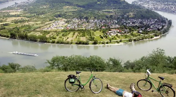 Pareja descansa junto a sus bicicletas en la ruta EuroVelo 15, con un río y un pueblo en el valle.