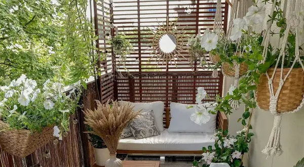 Przytulny balkon w bloku z drewnianymi zasłonami, sofą, białymi kwiatami i makramami.