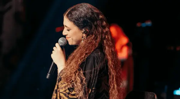 Cantante con lunghi capelli ricci, microfono in mano, sul palco. Un'espressione che evoca la bellezza della Sicilia, un testo che risuona.