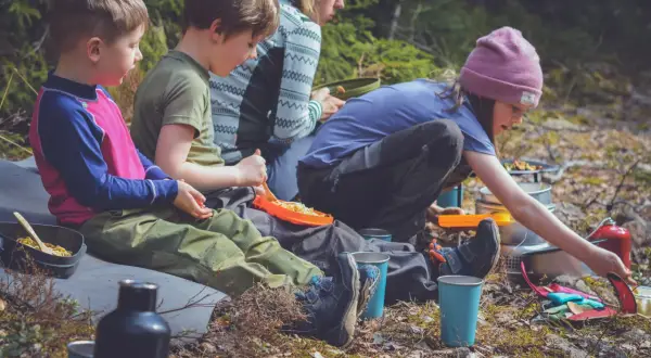 Familj njuter av en picknick i skogen, kanske vid restaurang Ånnaboda. Barn äter pasta och vuxna förbereder mat.