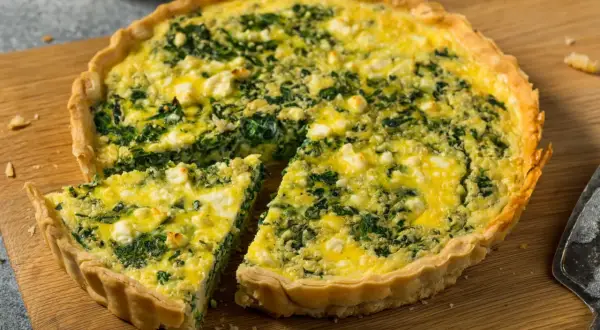 Pyszna tarta ze szpinakiem i serem feta, z jednym kawałkiem już wyciętym, prezentuje się apetycznie na drewnianej desce.