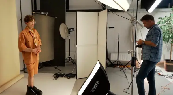 Model w pomarańczowym stroju pozuje w studiu, gdzie fotograf ustawia sprzęt, tworząc idealne naturalne źródła światła.