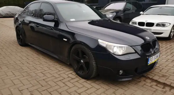 Ciemnogranatowe BMW E60 535d na czarnych felgach, zaparkowane na kostce brukowej przed budynkiem.