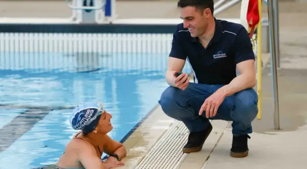 A swim coach crouches by the pool, holding a stopwatch, to discuss swim technique with a swimmer.