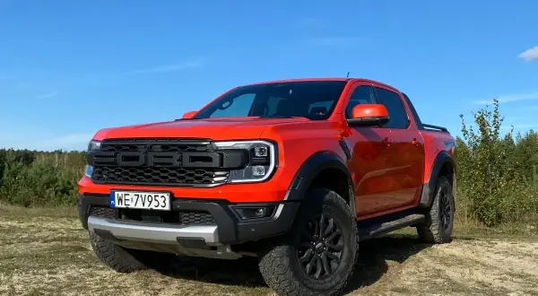 Pomarańczowy Ford Ranger Raptor gotowy na przygodę na piaszczystym terenie, z lasem w tle i błękitnym niebem.