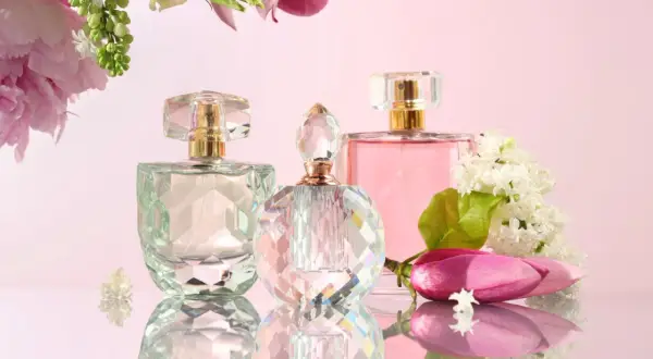 Trzy flakoniki perfum na różowym tle, otoczone kwiatami i liśćmi.