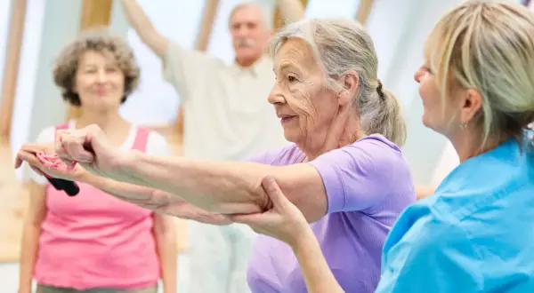 Seniorzy wykonują ćwiczenia na alzheimera z fizjoterapeutką. W tle inni uczestnicy ćwiczą z hantlami.