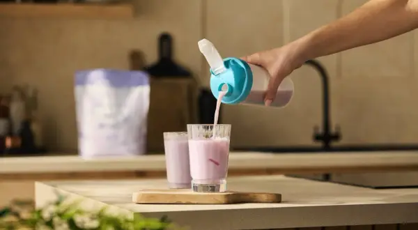 Eine Hand gießt einen rosa Proteinshake aus einem Shaker in ein Glas. So einfach ist es, was bewirken proteinshakes für die schnelle Nährstoffversorgung.