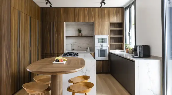 Cuisine moderne aux finitions boisées, conçue par un architecte d'intérieur. Table ovale en bois, tabourets, îlot central en marbre, et électroménagers intégrés.