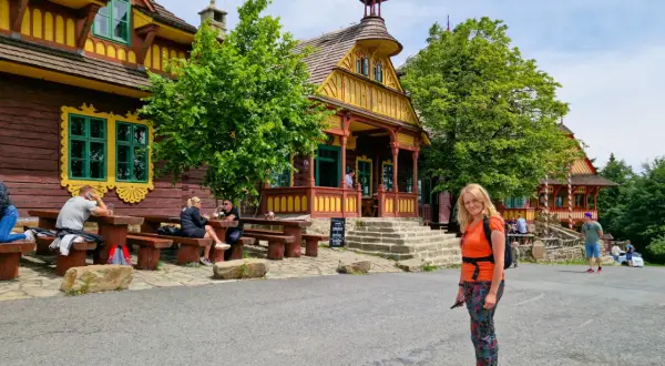 Kolorowe, drewniane budynki w Czechach, pełne ciekawostek dla dzieci. Kobieta w pomarańczowej bluzce pozuje na tle malowniczej architektury.