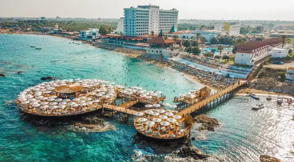 Idealne wakacje na Cyprze: turkusowe morze, plaża z leżakami i parasolami, hotel i restauracja na wodzie.