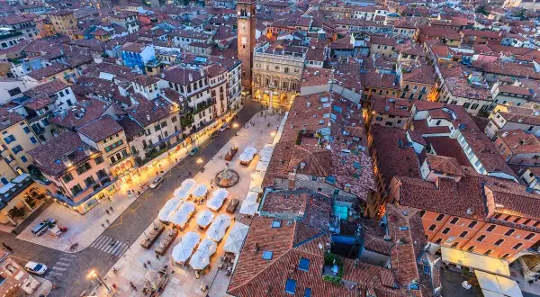 Vista aerea di piazza Erbe a Verona, con i suoi edifici storici, la torre e i tavolini all'aperto.