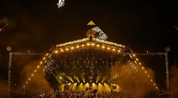A performer on a bicycle is suspended in the air above a brightly lit stage at Glastonbury. The crowd below anticipates the next act, checking their Glastonbury set times.