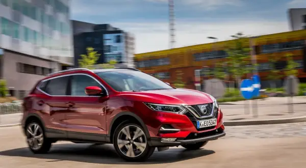 Czerwony Nissan Qashqai 1 jedzie ulicą w mieście. W tle nowoczesne budynki i niebieski znak drogowy.
