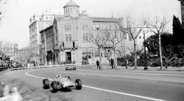 Coches de carreras compiten en el circuito de Montjuïc, con espectadores en las gradas y edificios históricos al fondo.