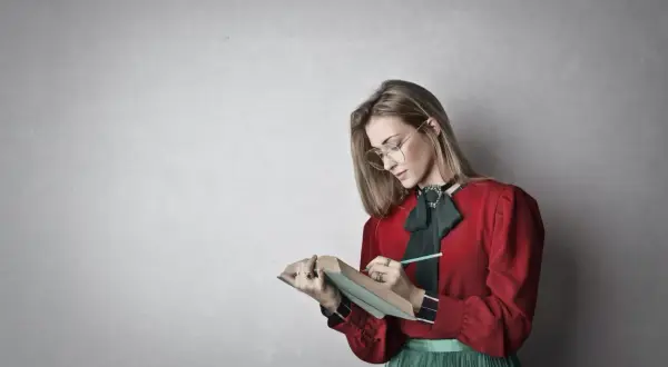 Młoda kobieta w okularach i czerwonej bluzce z zieloną kokardą pochyla się nad książką, notując coś ołówkiem. To inspiracja, jak napisać esej historyczny.