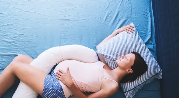 Mujer embarazada durmiendo de lado con almohadas de apoyo, una solución para como dormir con pubalgia y disfrutar del descanso.