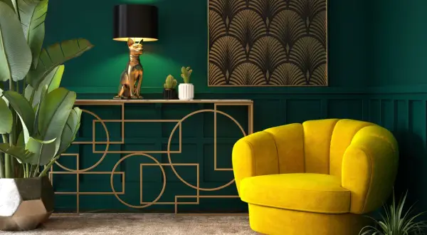Elegancka tkanina art deco zdobi ścianę nad złotą konsolą z lampą w kształcie kota. Obok żółty fotel i bujna roślina.