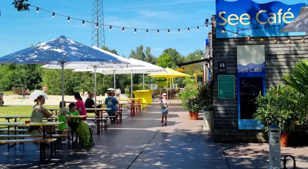 See Café mit Tischen und Gästen unter Sonnenschirmen am See.