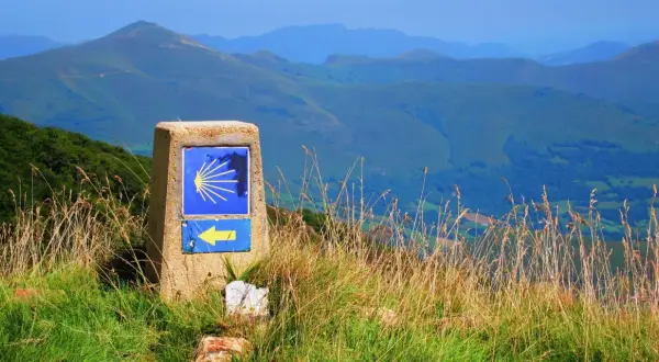 Kamień z symbolem muszli i żółtą strzałką, wskazujący drogę na Santiago de Compostela szlak z Polski. W tle góry.