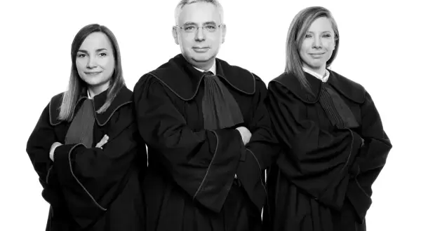 Trzy osoby w togach prawniczych, dwie kobiety i mężczyzna, stoją na białym tle.