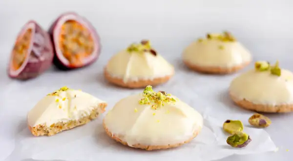 Små, vita bakelser med pistagecrunch, inspirerade av passionsfrukt recept. En är uppskuren och visar en krämig fyllning.