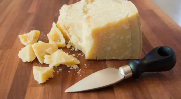 Kawałek parmezanu i kilka mniejszych kawałków na desce. Obok leży nóż do sera. Warto sprawdzić parmezan kcal przed spożyciem.