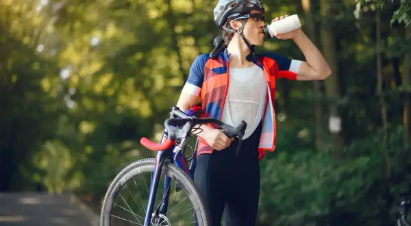 Kolarz w stroju sportowym pije wodę z bidonu, dbając o odpowiednią dietę kolarza. W tle zielony las.
