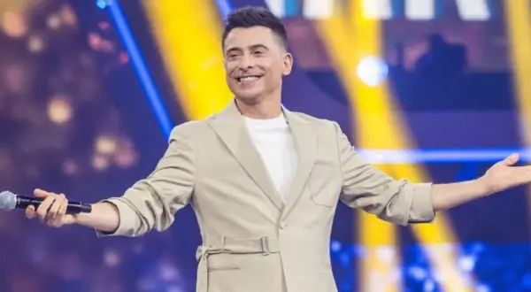 Uśmiechnięty mężczyzna w beżowej marynarce trzyma mikrofon, stoi na tle kolorowej sceny.