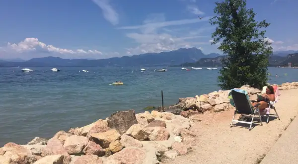 Una donna legge su una sedia a sdraio sulla riva rocciosa di Cisano, Gardasee. Barche galleggiano sull'acqua calma con montagne sullo sfondo.