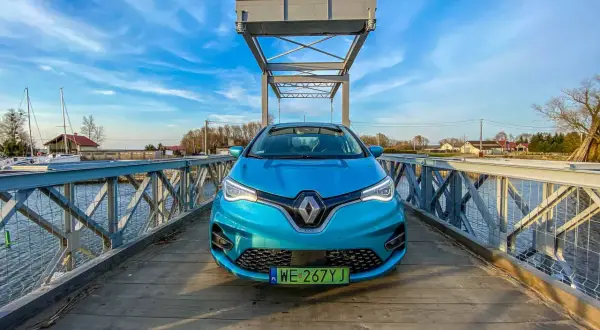 Niebieski Renault Zoe stoi na drewnianym moście nad wodą, z żaglówkami w tle.