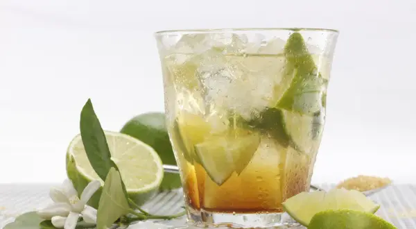 Orzeźwiająca caipirinha przepis: drink z lodem, limonką i cukrem. Obok leżą świeże limonki i kwiatki.