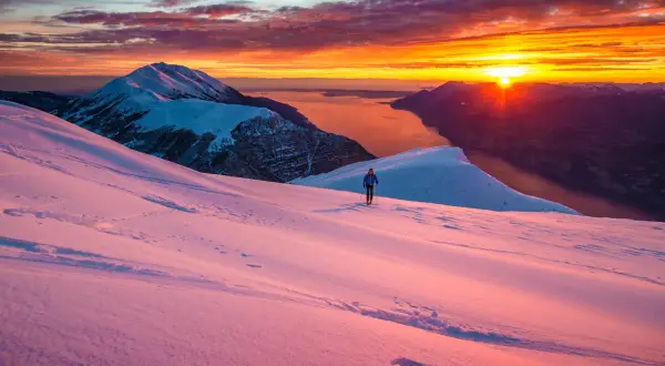 Alba infuocata sul lago di Garda in inverno. Un escursionista solitario cammina su un pendio innevato, godendosi la vista mozzafiato.