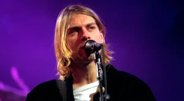 Kurt Cobain śpiewa do mikrofonu na tle fioletowej sceny.