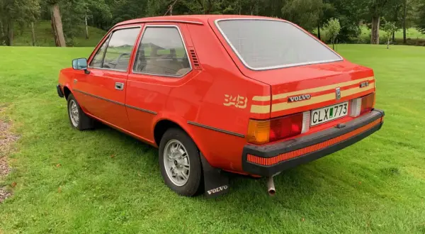 En röd Volvo 343 står på en grön gräsmatta med skog i bakgrunden. Bilen har en gul rand längs sidan och texten "343" i guld.