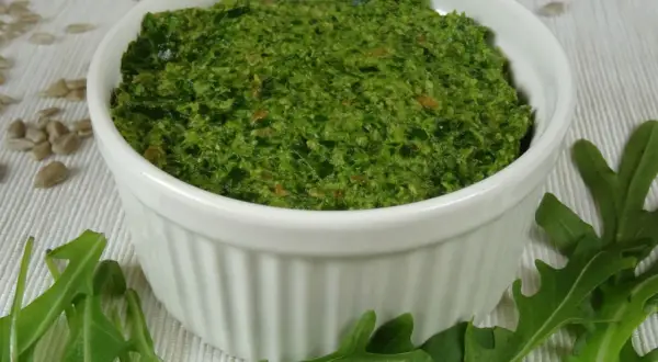 Zielone pesto z rukoli w białej miseczce, otoczone liśćmi rukoli i nasionami słonecznika.
