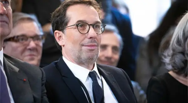 Un homme avec des lunettes, en costume noir, souriant.