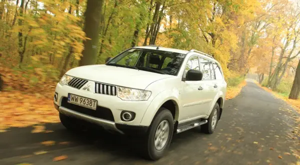 Biały model terenówki Mitsubishi Pajero Sport jedzie po drodze wśród jesiennych drzew.