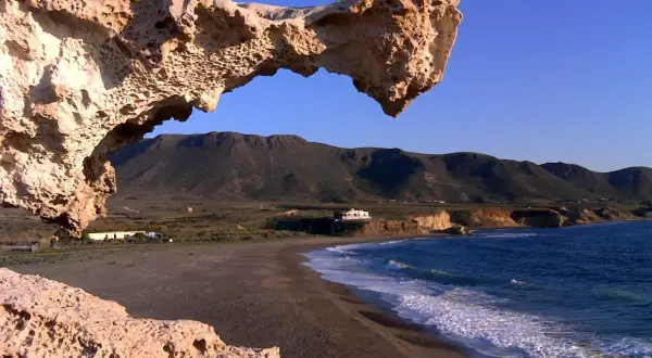 Un arco natural de roca erosionada, como un gigante dormido, vigila una playa solitaria y el mar azul. Los escullos forman un portal a un paisaje sereno.