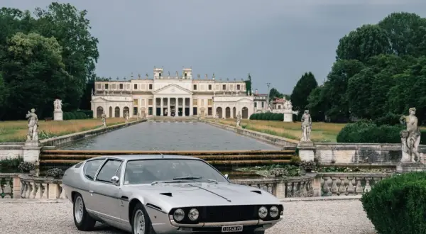Srebrny Lamborghini Espada przed okazałą willą z basenem i rzeźbami. Klasyk motoryzacji w malowniczej scenerii.