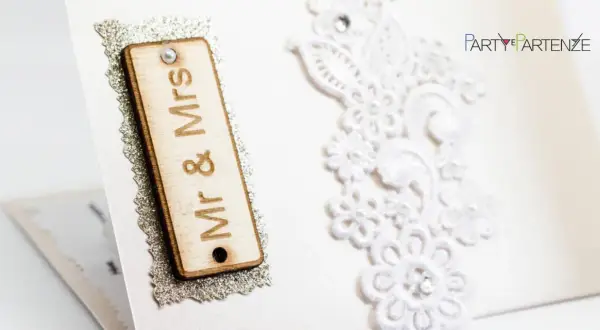 Dettagli di un invito di nozze: targa in legno "Mr & Mrs", pizzo bianco e logo "Party Partenze". Un esempio di diversi tipi di nozze.